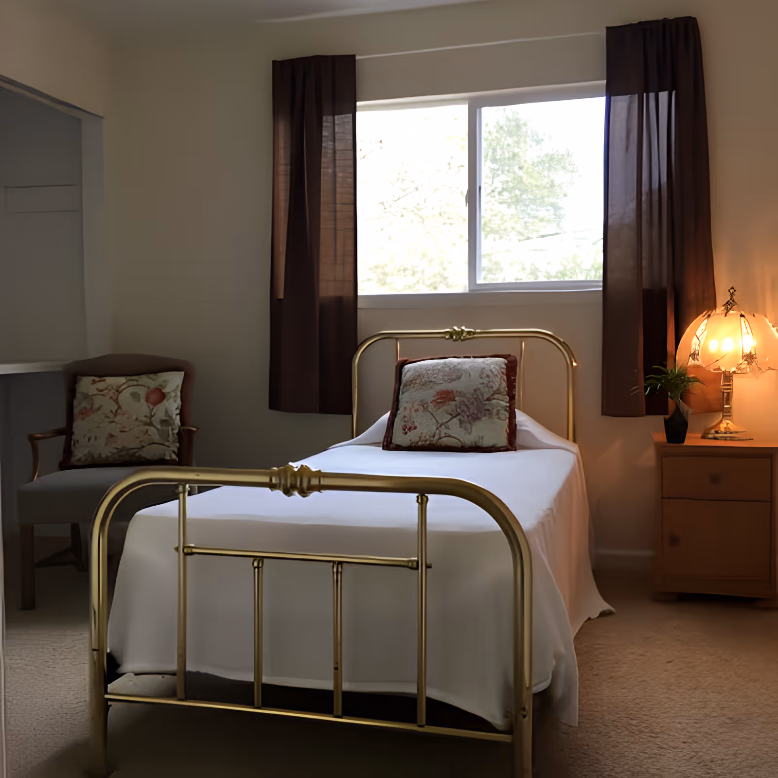 A small bedroom with a brass bed frame, a white bedspread, and a decorative pillow. There is a window with dark curtains behind the bed, a wooden nightstand with a vintage lamp and a small plant, and an upholstered chair with a floral cushion in the corner.