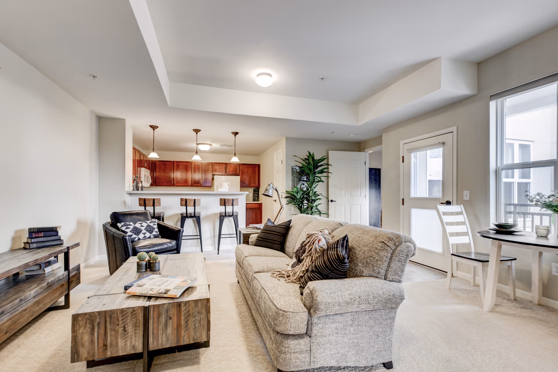 A bright and spacious living room area in a senior living facility with a beige sofa adorned with pillows and a throw blanket, a wooden coffee table with magazines and small plants, a black armchair with a patterned cushion, and a wooden TV stand with books. In the background, there is a kitchen with a breakfast bar and three bar stools, pendant lights hanging from the ceiling, and wooden cabinets. To the right, there is a small dining table with two chairs near a window and a door leading outside. The room has light-colored walls and carpeted floors.