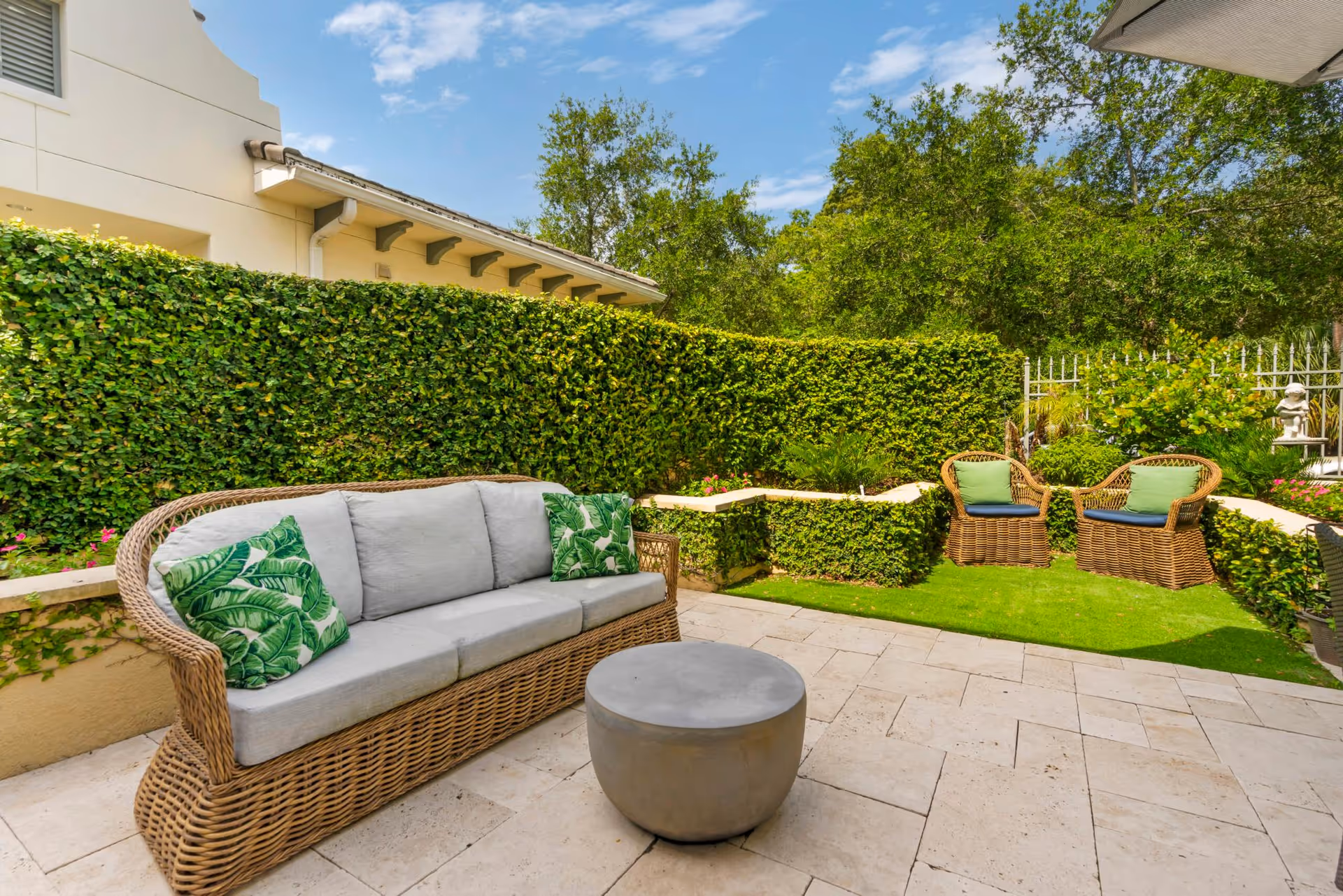 Outdoor patio area with a wicker sofa featuring gray cushions and green leaf-patterned pillows, two wicker chairs with green cushions, a round gray coffee table, surrounded by lush green hedges and trees under a blue sky.