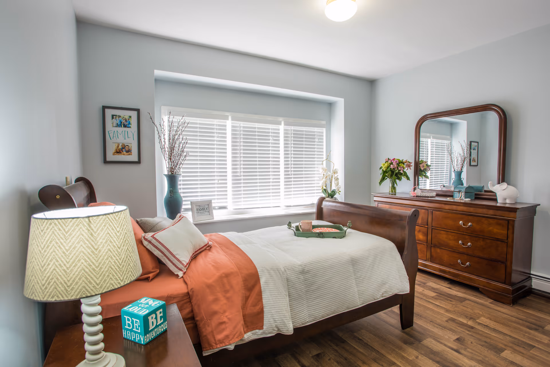 Sunlit bedroom with a wooden sleigh bed, orange and white bedding, a dresser with mirror, bedside table and lamp, and a window with blinds.
