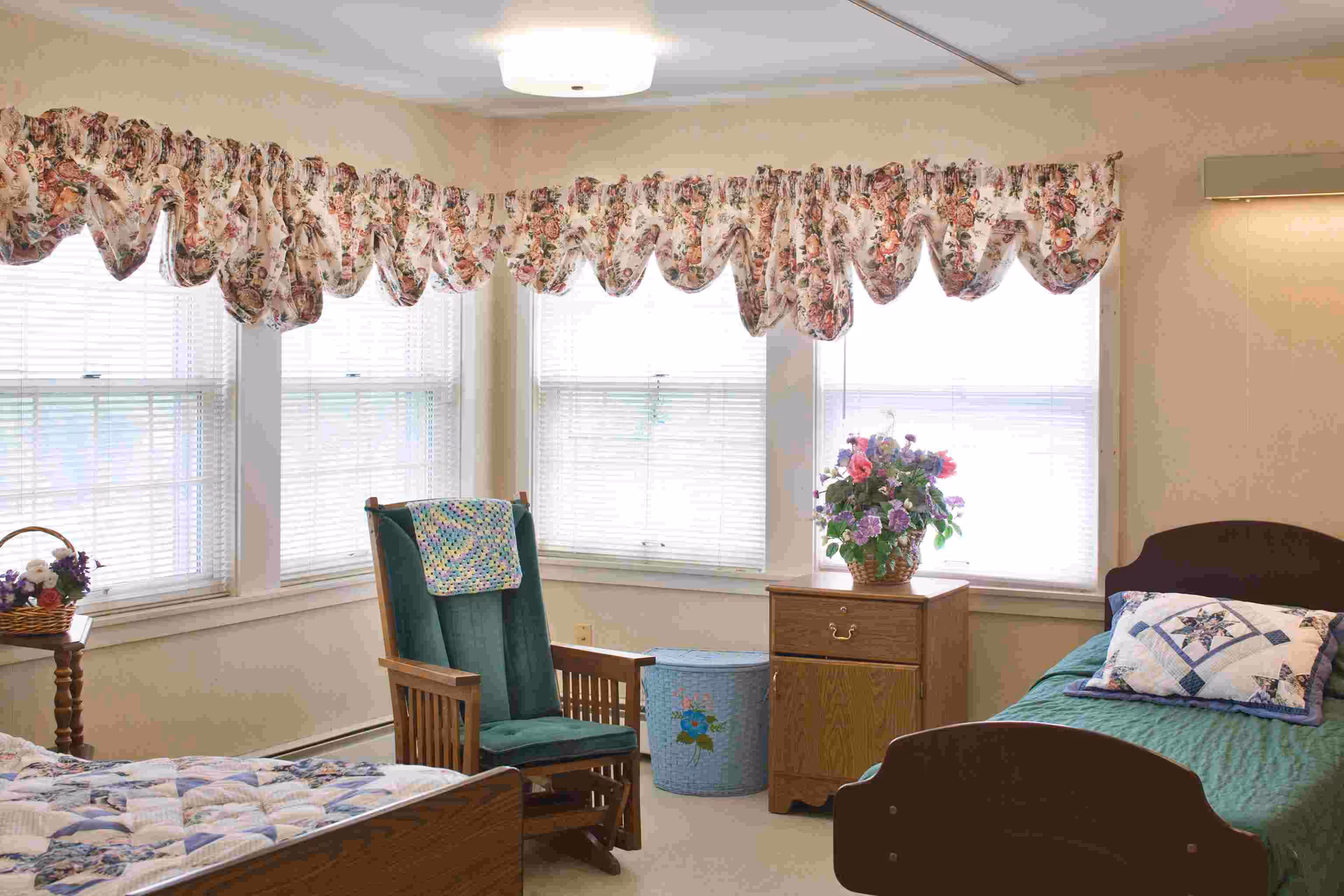 A cozy bedroom with two single beds, each with a quilted bedspread and decorative pillows. Between the beds is a wooden nightstand with a basket of flowers on top. A green cushioned rocking chair with a crocheted blanket draped over the back sits near a corner with three windows covered by floral valances. A blue laundry basket with a flower design is next to the nightstand. The room has soft beige walls and is well-lit by natural light from the windows.