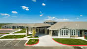 Exterior view of a single-story assisted living facility with a covered entrance, large windows, and a well-maintained lawn under a blue sky with scattered clouds.
