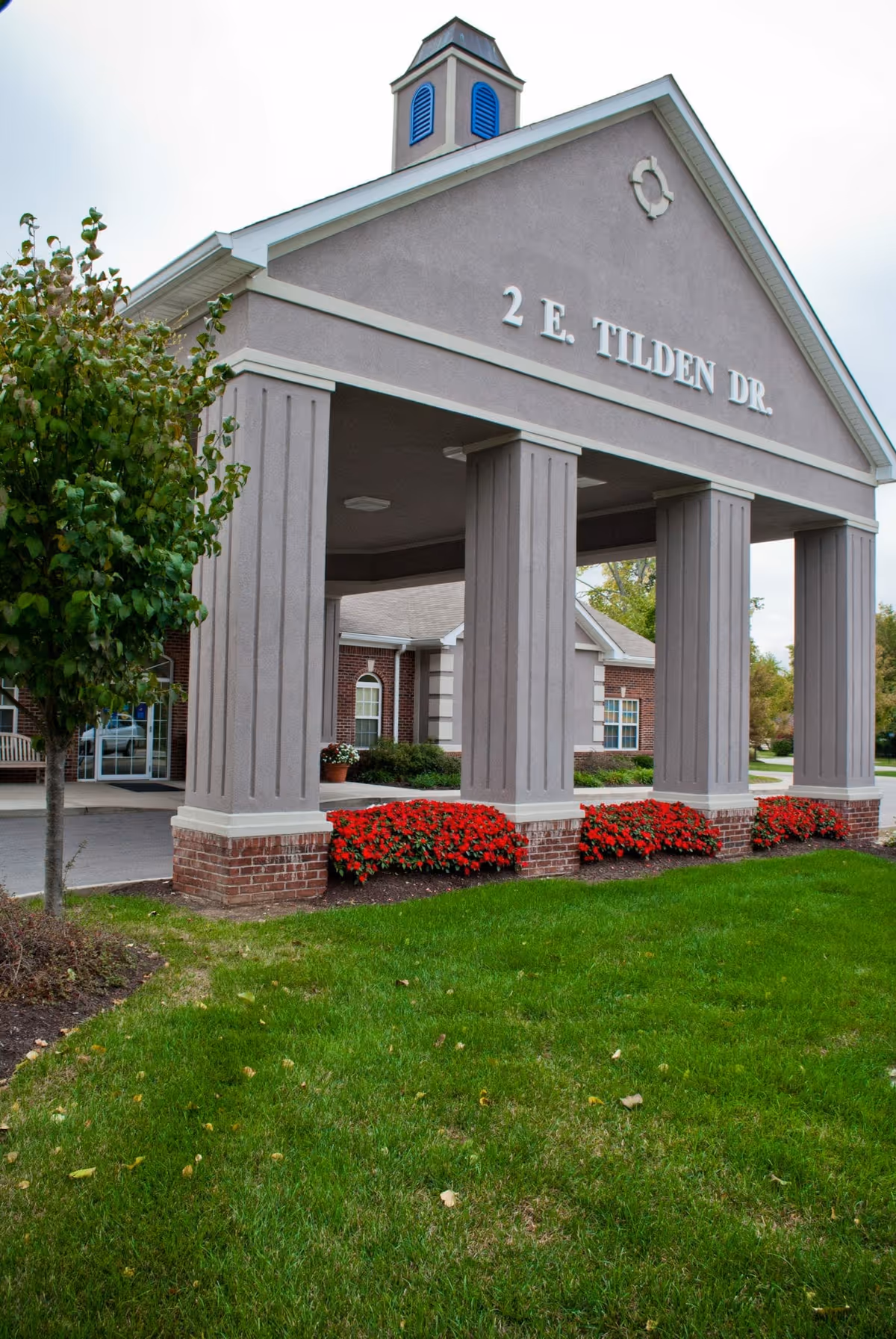 Entrance of a building with a covered driveway supported by four large columns. The address '2 E. TILDEN DR.' is displayed prominently on the front of the structure. There are red flowers planted along the base of the columns and a small tree to the left. The building has a brick and gray exterior with a small cupola on top.
