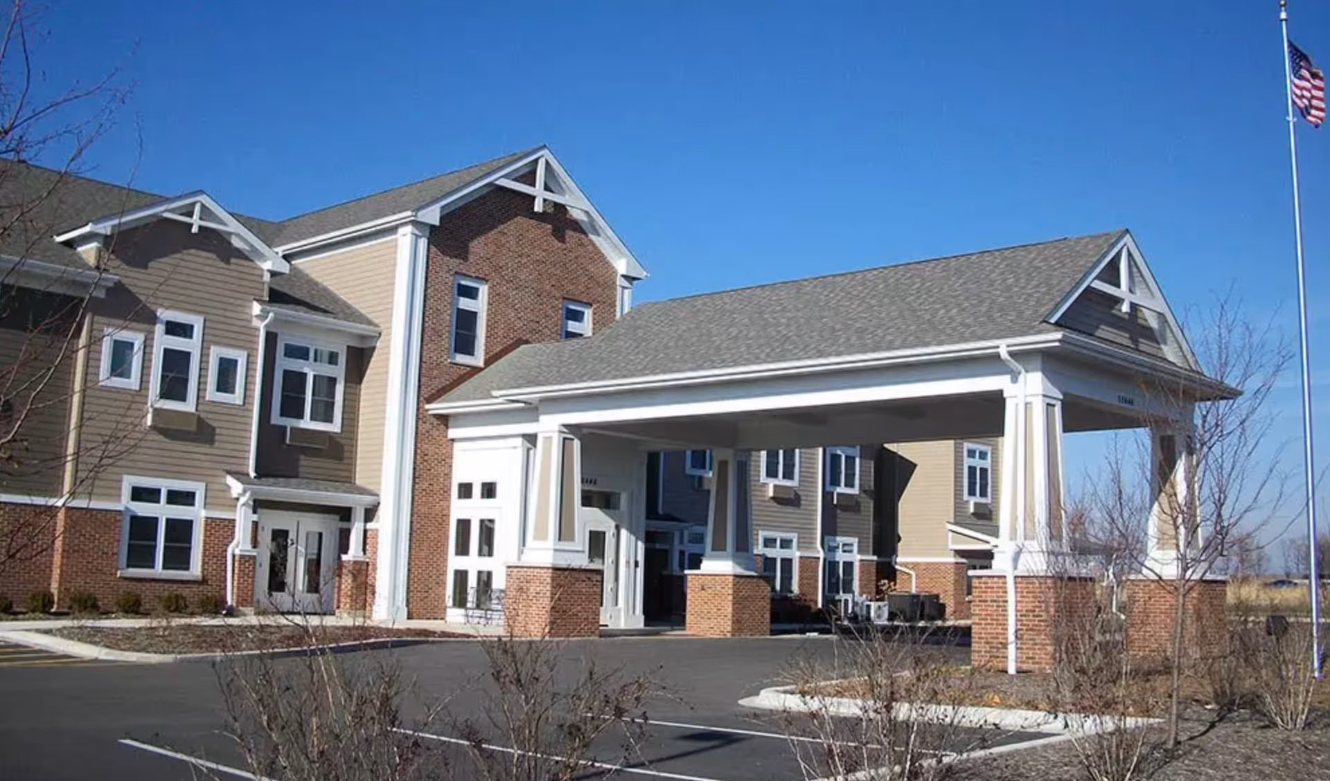 Front exterior of a multi-story senior living building with a covered porte-cochère, brick-and-siding facade, and an American flag against a clear blue sky.