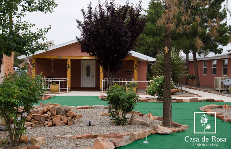 Exterior view of Casa de Rosa Assisted Living showing a single-story building with a front porch, surrounded by trees, bushes, and a landscaped area with rocks and a small putting green with golf flags.