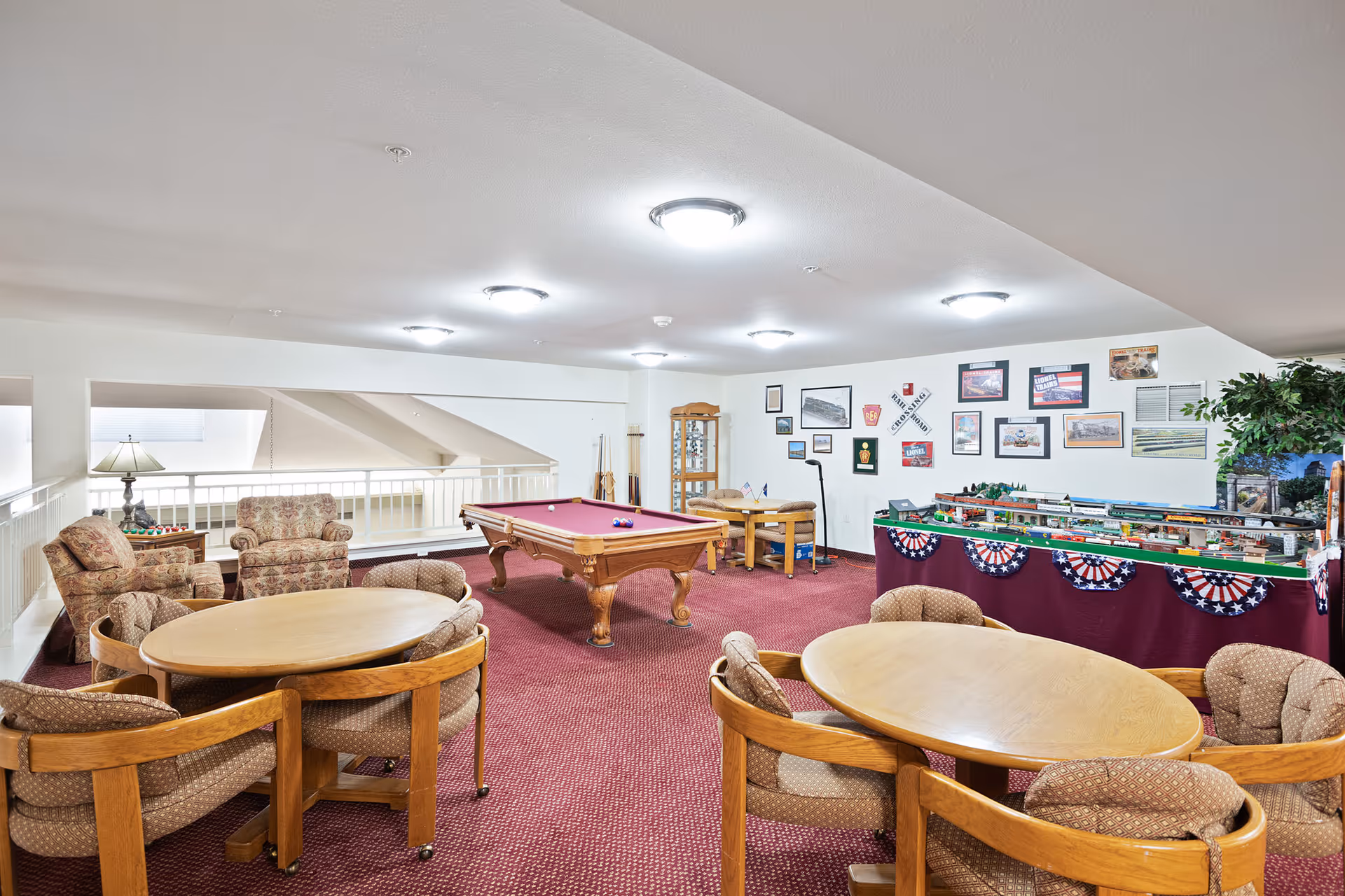 A recreational room with a red carpet featuring a pool table in the center, two round wooden tables each surrounded by four cushioned chairs, two upholstered armchairs near a lamp, and a display of model trains and framed pictures on the wall.