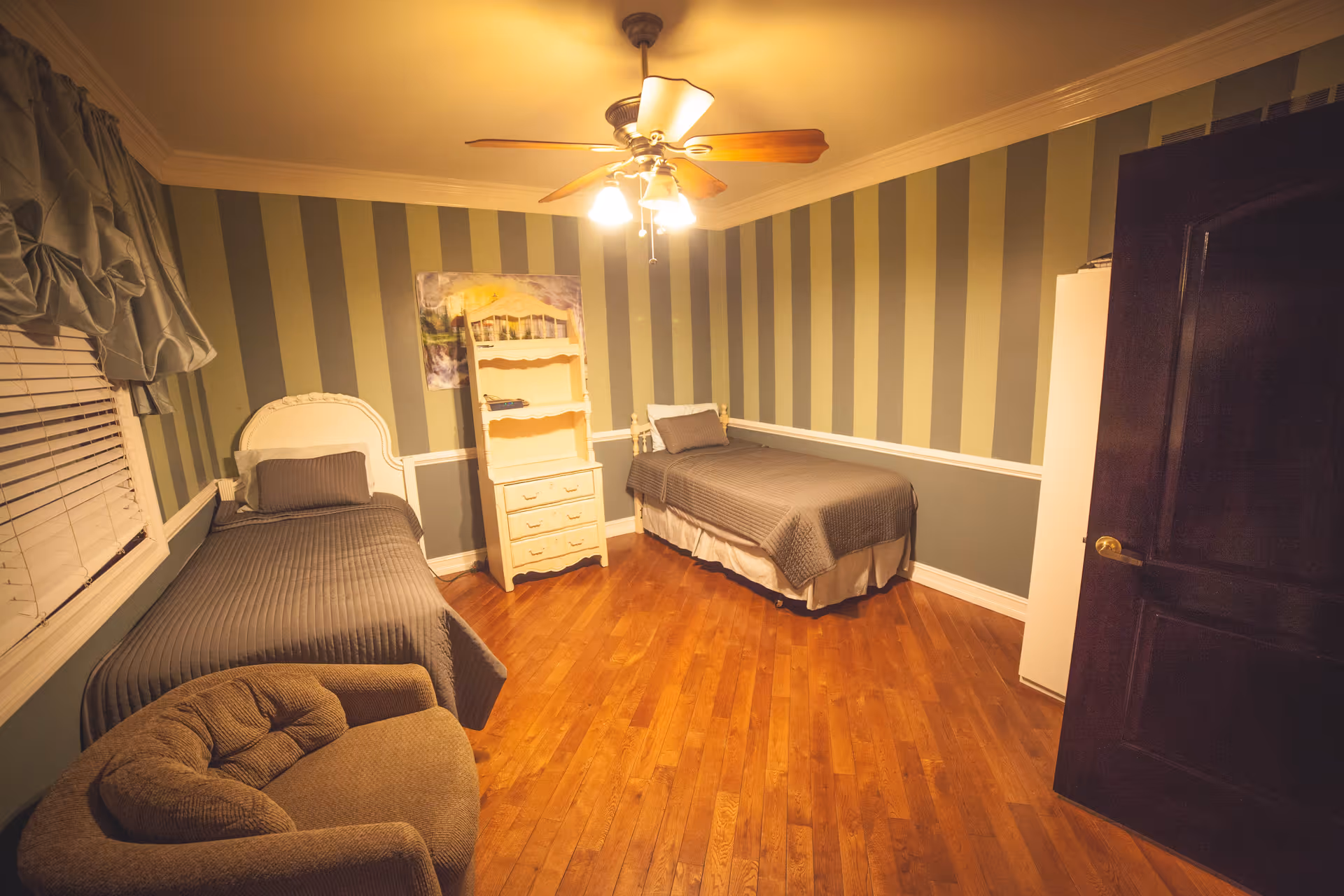 A bedroom with two single beds covered in gray bedspreads, a beige armchair, a white dresser with shelves, striped green and gray walls, a ceiling fan with lights, and hardwood flooring.