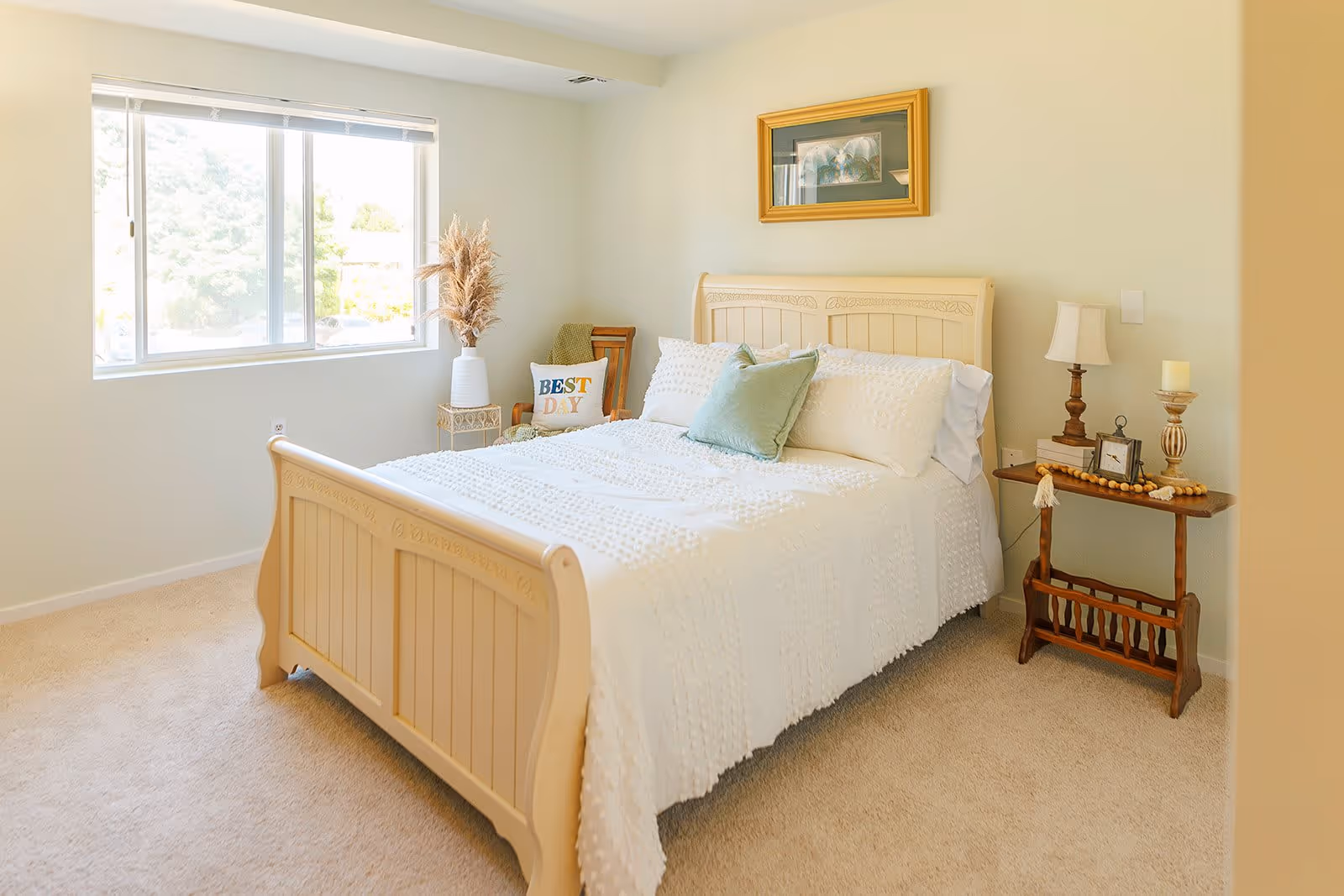 A bright bedroom with a large window letting in natural light. The room features a cream-colored wooden bed with white bedding and decorative pillows, a wooden side table with a lamp, clock, and candle, a chair with a cushion that says 'BEST DAY', and a framed picture hanging on the wall above the bed.
