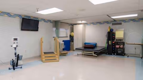 An empty therapy room with exercise equipment, stairs, mats, and a wall-mounted TV.