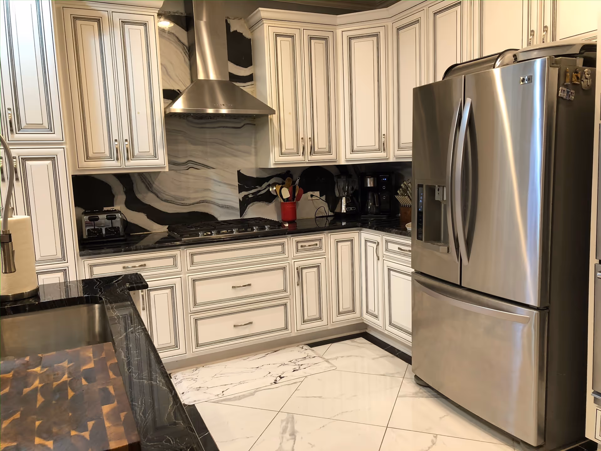 Modern kitchen with white cabinetry, black marble countertops, a stainless steel refrigerator, a gas stove with a stainless steel range hood, a coffee maker, toaster, and various kitchen utensils on the counter. The floor is tiled with white marble tiles.