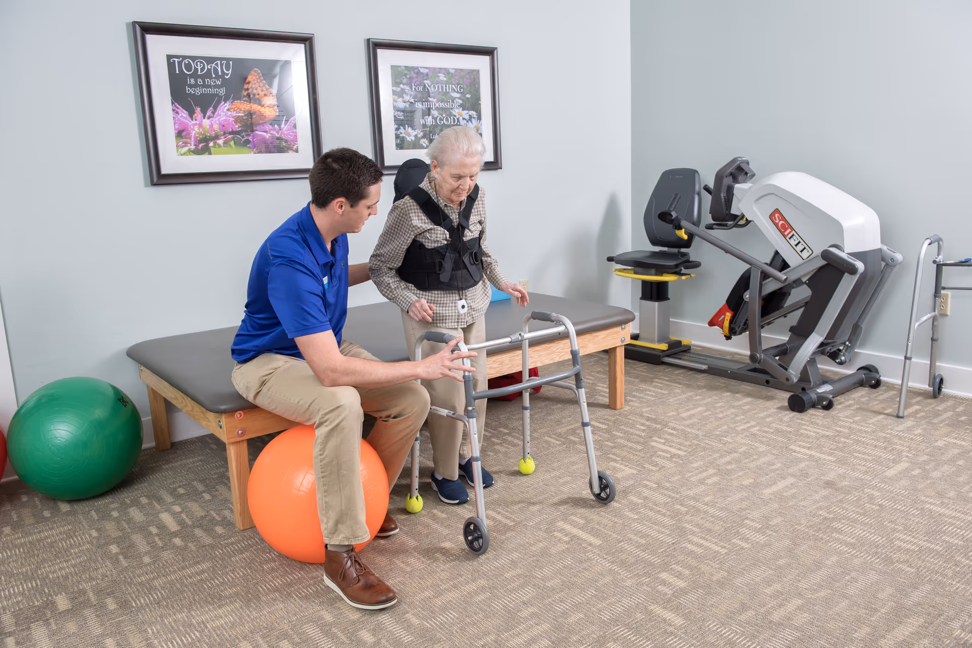 A caregiver assists an elderly woman using a walker in a rehabilitation/exercise room with fitness equipment and exercise balls.