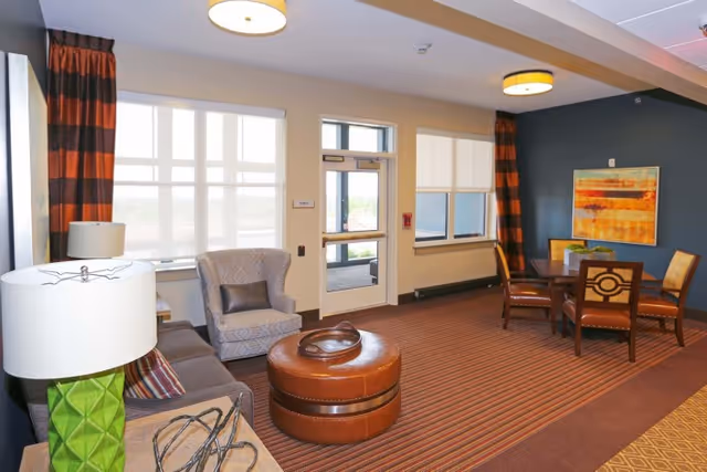 A cozy living room area with a striped carpet, a gray armchair with a pillow, a brown leather ottoman, a wooden dining table with four chairs, and a large abstract painting on the dark blue wall. The room has large windows with white blinds and brown striped curtains, and ceiling lights providing warm illumination.