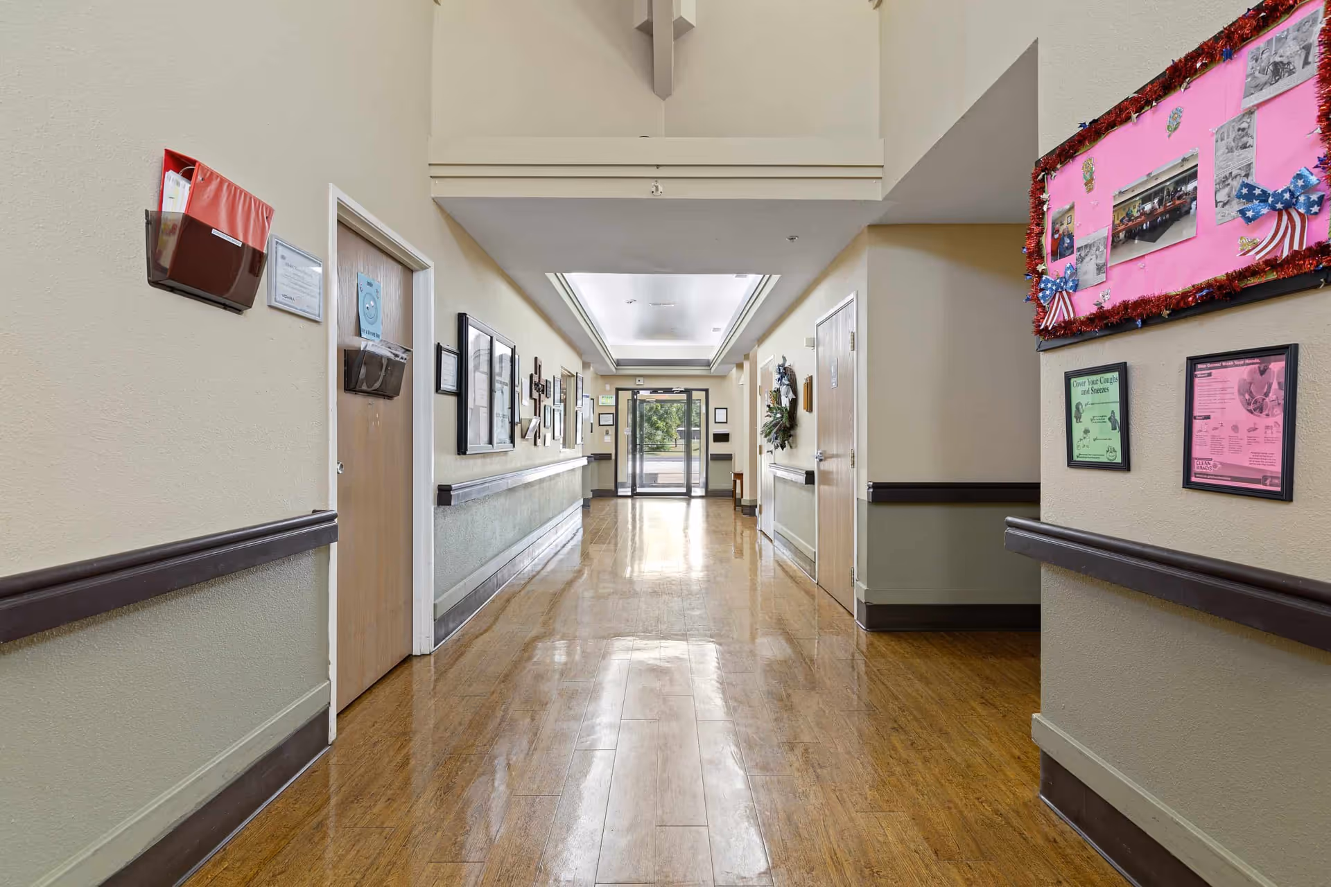 A clean, well-lit hallway in a nursing and rehab facility with wooden floors, beige walls, handrails on both sides, bulletin boards with notices and decorations, and a glass exit door at the far end.