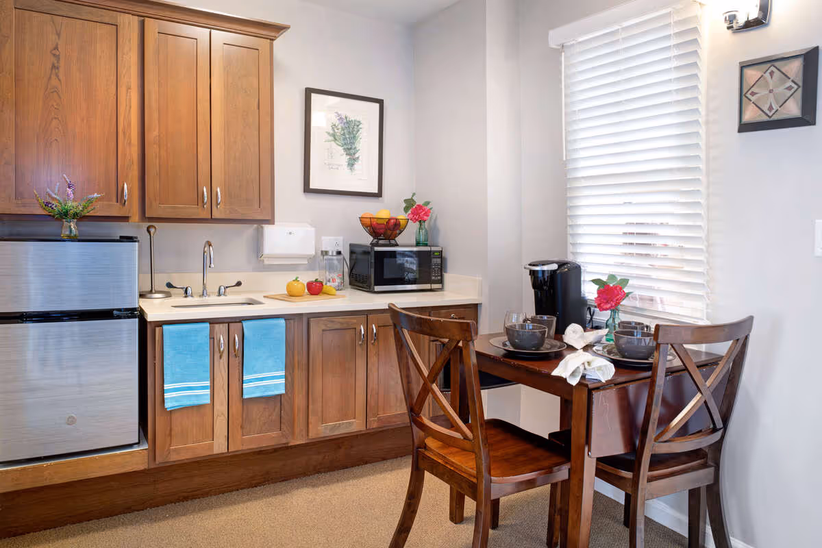 Small kitchenette with wooden cabinets, a sink, mini-fridge, microwave, and a two-person dining table set by a window.