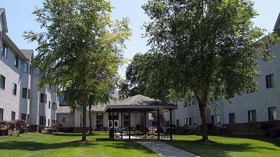 Outdoor courtyard with a gazebo and trees between two wings of a multi-story senior living building.