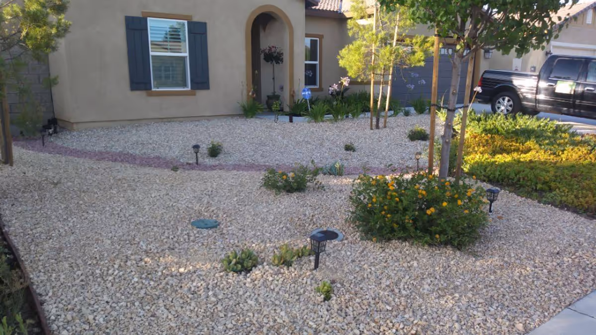 Front yard of a house with a gravel landscape, small plants, and a few young trees. The house has beige walls, a window with blue shutters, and an arched doorway. A black pickup truck is parked in the driveway to the right.