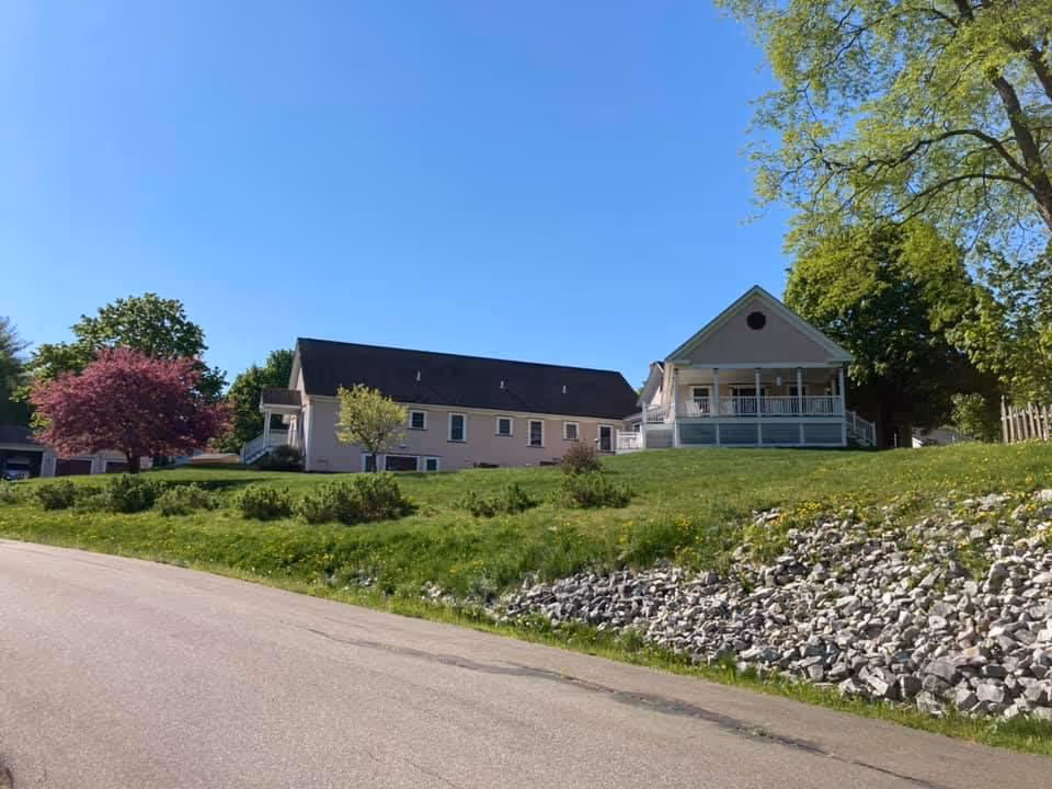 Light-colored multi-section building with a covered porch on a grassy hill, trees, and a clear blue sky.