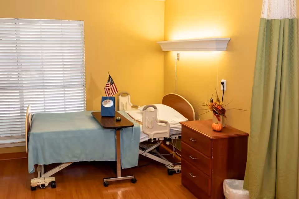 A hospital-style bed with blue linens in a yellow-walled patient room beside a wooden bedside cabinet, window blinds, and a green privacy curtain.