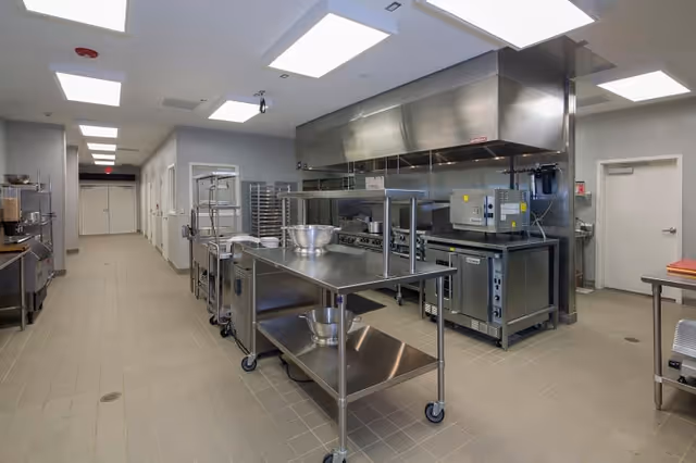 A clean, modern commercial kitchen with stainless steel work tables, cooking appliances, and ventilation hoods. The kitchen has tiled floors, bright overhead lighting, and various kitchen equipment including mixing bowls and racks.