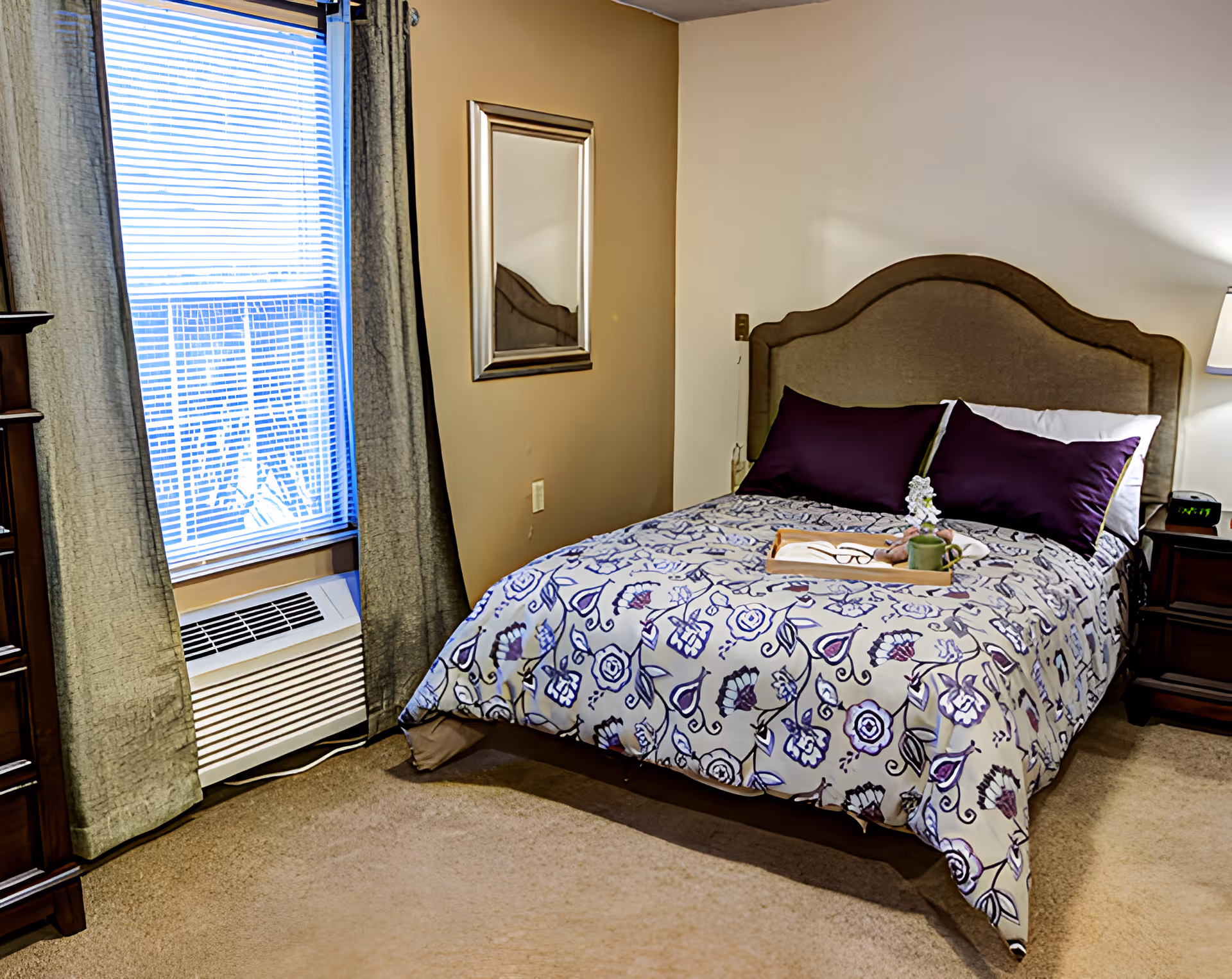 Bedroom with a bed dressed in a floral comforter, upholstered headboard, nightstand with lamp, and a curtained window.