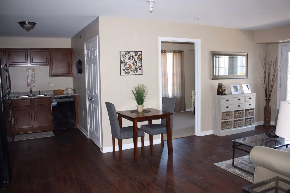 Interior view of a senior living facility showing a small dining area with a wooden table and two gray chairs, a kitchen with wooden cabinets and black appliances on the left, and a living room area with a white cabinet, decorative items, and a glass coffee table on the right. The walls are beige, and the floor is dark wood. A doorway leads to another room with curtains.