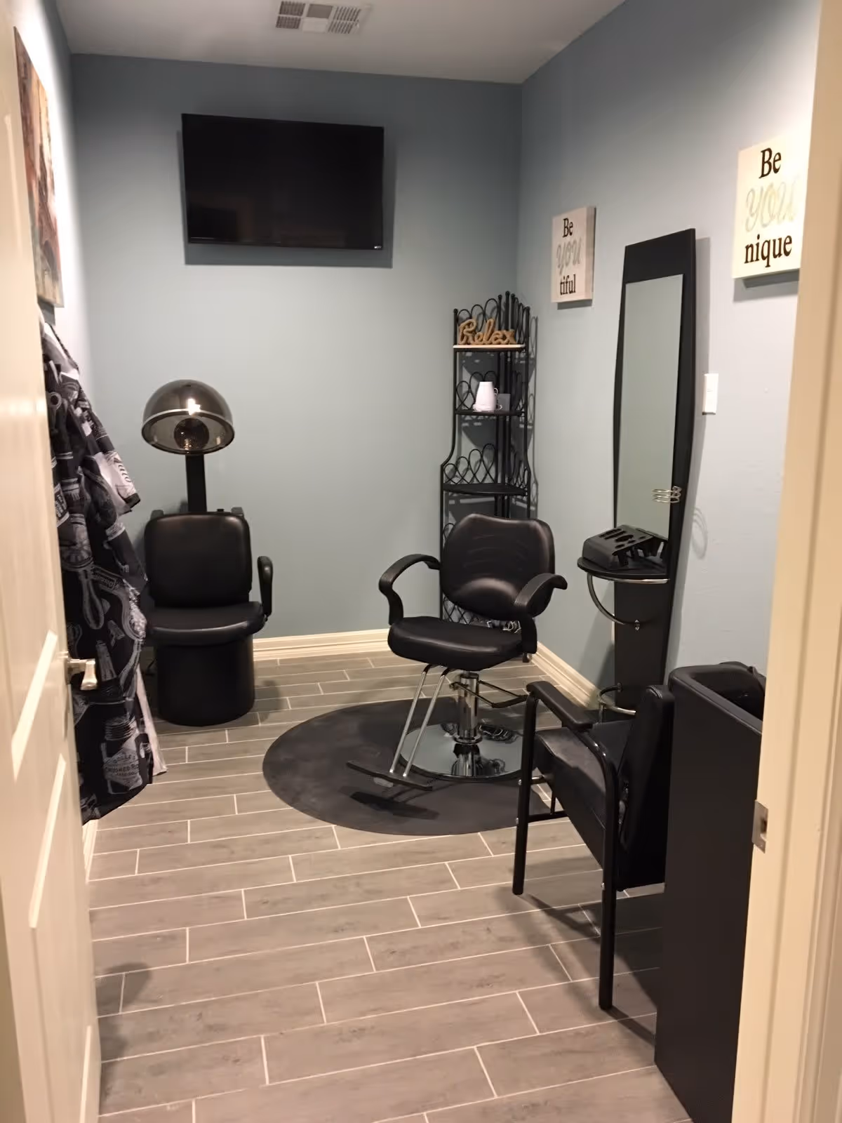 Interior view of a small salon room with a black salon chair on a circular mat, a hair dryer chair, a wall-mounted TV, a black metal shelf with decorative items, a mirror, and wall art with motivational quotes on light blue walls and gray tiled floor.