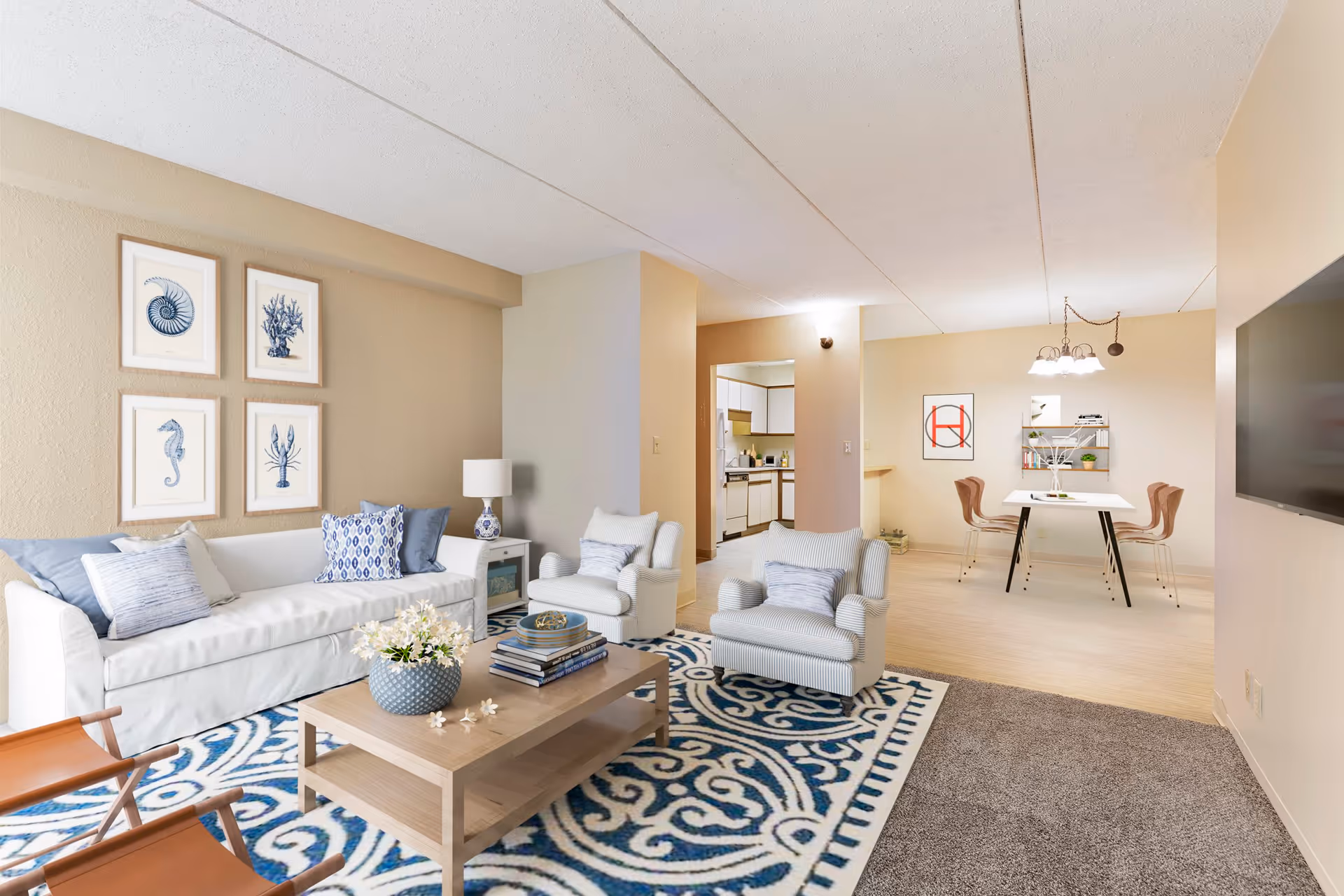 A cozy living room area with a white sofa adorned with blue and white pillows, two striped armchairs, a wooden coffee table with a vase of flowers and books, and a blue and white patterned rug. The room has beige walls with four framed sea-themed artworks. In the background, there is a dining area with a white table, four chairs, a hanging light fixture, and a small kitchen visible through an open doorway.