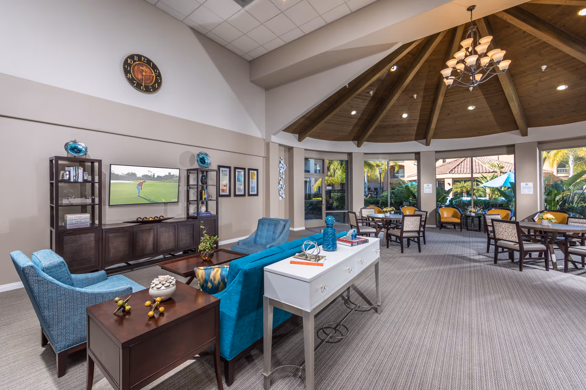 A spacious and well-lit common area in a senior living facility featuring a vaulted wooden ceiling with a chandelier, a seating area with blue upholstered chairs and a sofa, a wall-mounted TV, decorative shelves, and several tables and chairs near large windows overlooking a garden with palm trees and umbrellas.