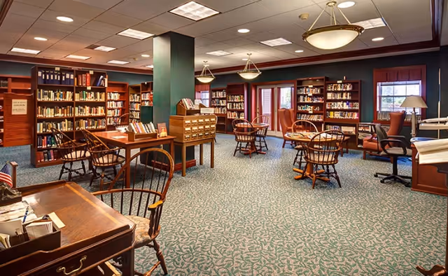 A spacious library room with multiple wooden bookshelves filled with books, several round wooden tables with chairs, a card catalog cabinet, and large windows letting in natural light. The room has a patterned carpet and ceiling lights.