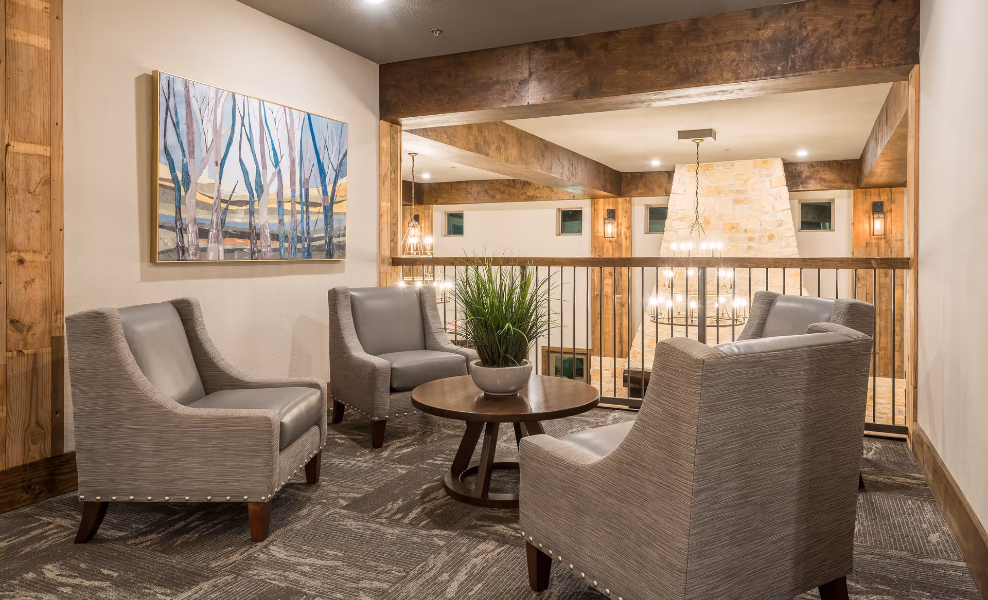 A cozy seating area with four gray upholstered armchairs arranged around a round wooden table with a potted plant on top. The space features wooden beams, a stone fireplace in the background, and a large painting of trees on the wall.