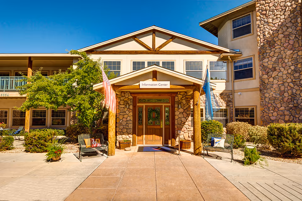 Front entrance of a senior living facility named The Chateau at Gardnerville with a stone and wood exterior, an 'Information Center' sign above the door, two flags on either side of the entrance, and outdoor seating with decorative pillows.