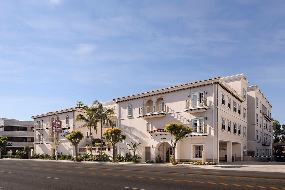 Exterior view of Belmont Village Senior Living Encino, a multi-story building with balconies, palm trees, and a clear blue sky.