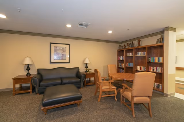 A cozy common area with a black leather couch and matching ottoman, two wooden side tables each with a lamp, a round wooden table surrounded by four cushioned chairs, and a wooden bookshelf filled with books and decorative items against a beige wall.