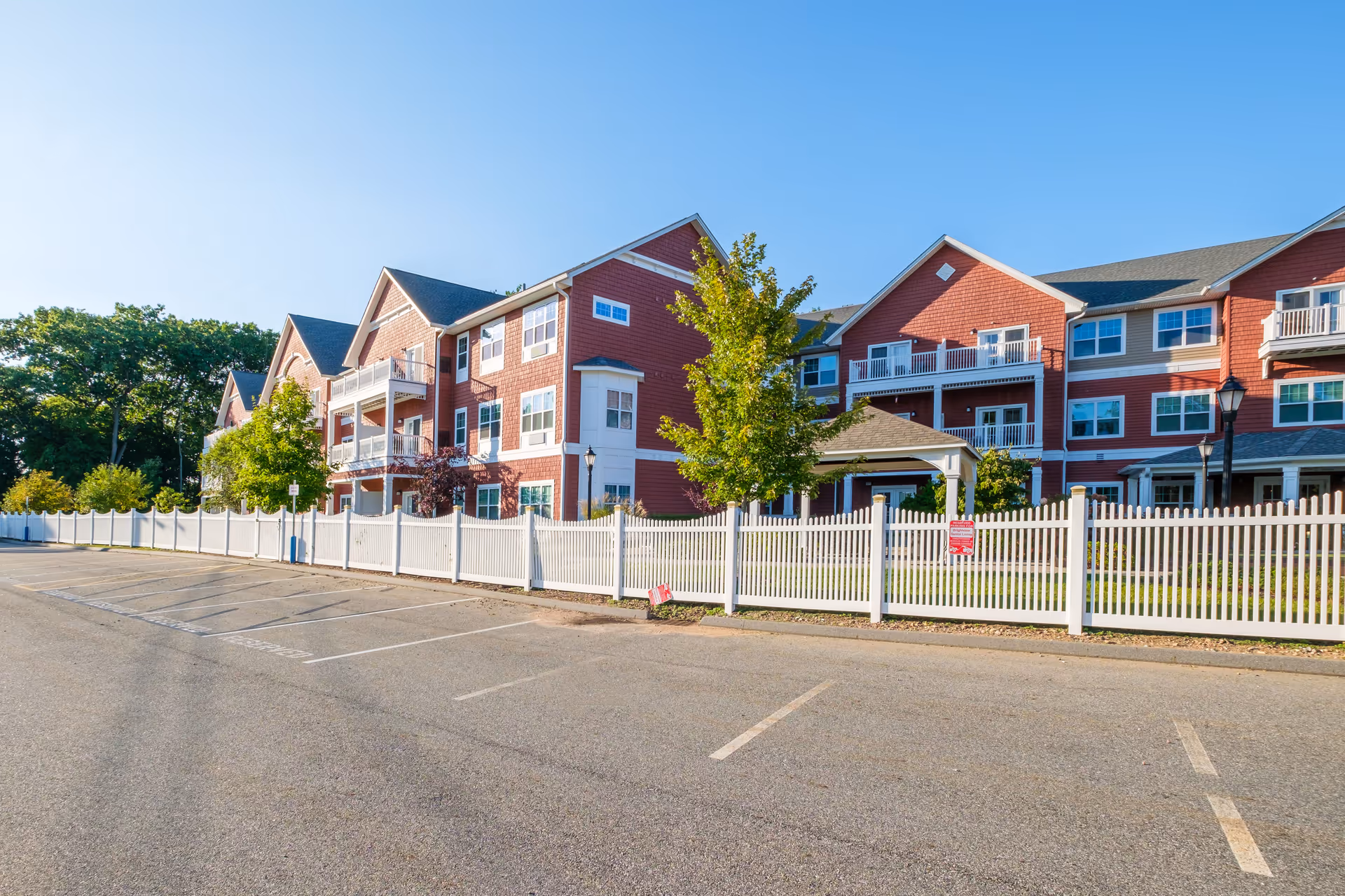 Exterior view of a senior living facility with red siding and white trim, multiple balconies, and a white picket fence surrounding a grassy area with trees and a small gazebo. The sky is clear and blue, and there is an empty parking lot in the foreground.