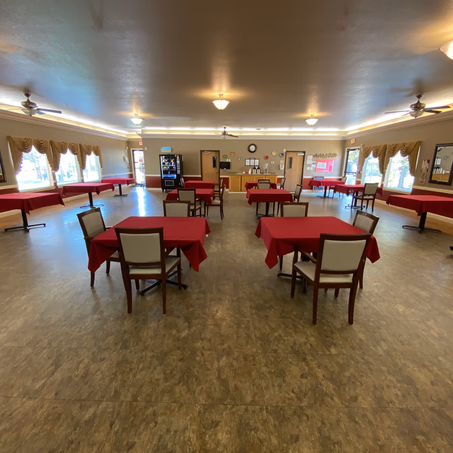 Large dining room with multiple tables covered in red tablecloths and chairs, windows with drapes, and a vending/serving area along the far wall.