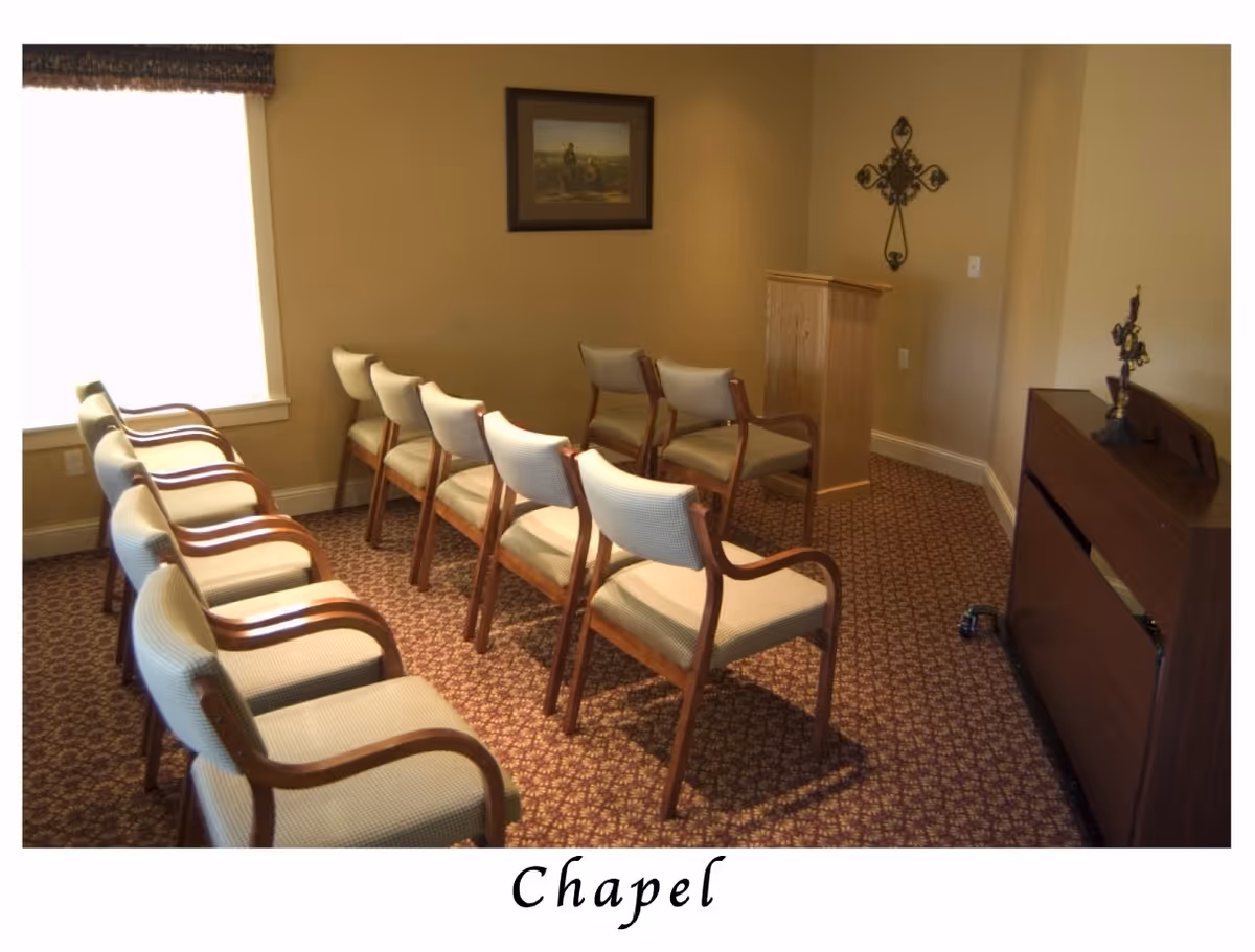 Small chapel room with rows of upholstered chairs facing a wooden lectern and decorative cross.
