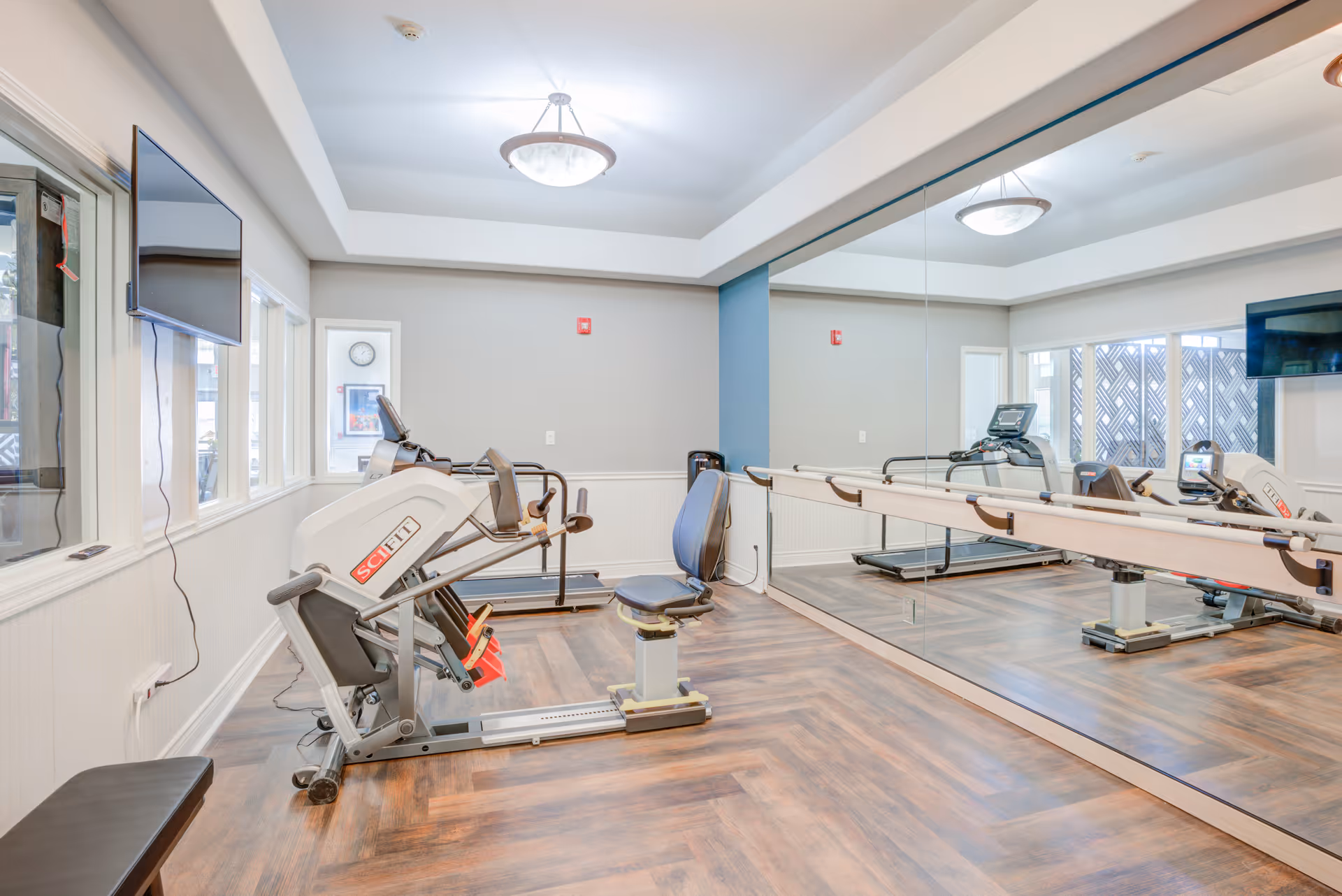 A bright exercise room with wood flooring, featuring a seated leg press machine and a treadmill. The room has large mirrors on one wall, a mounted flat-screen TV, and windows letting in natural light. The walls are painted light gray with white wainscoting and a blue accent column.
