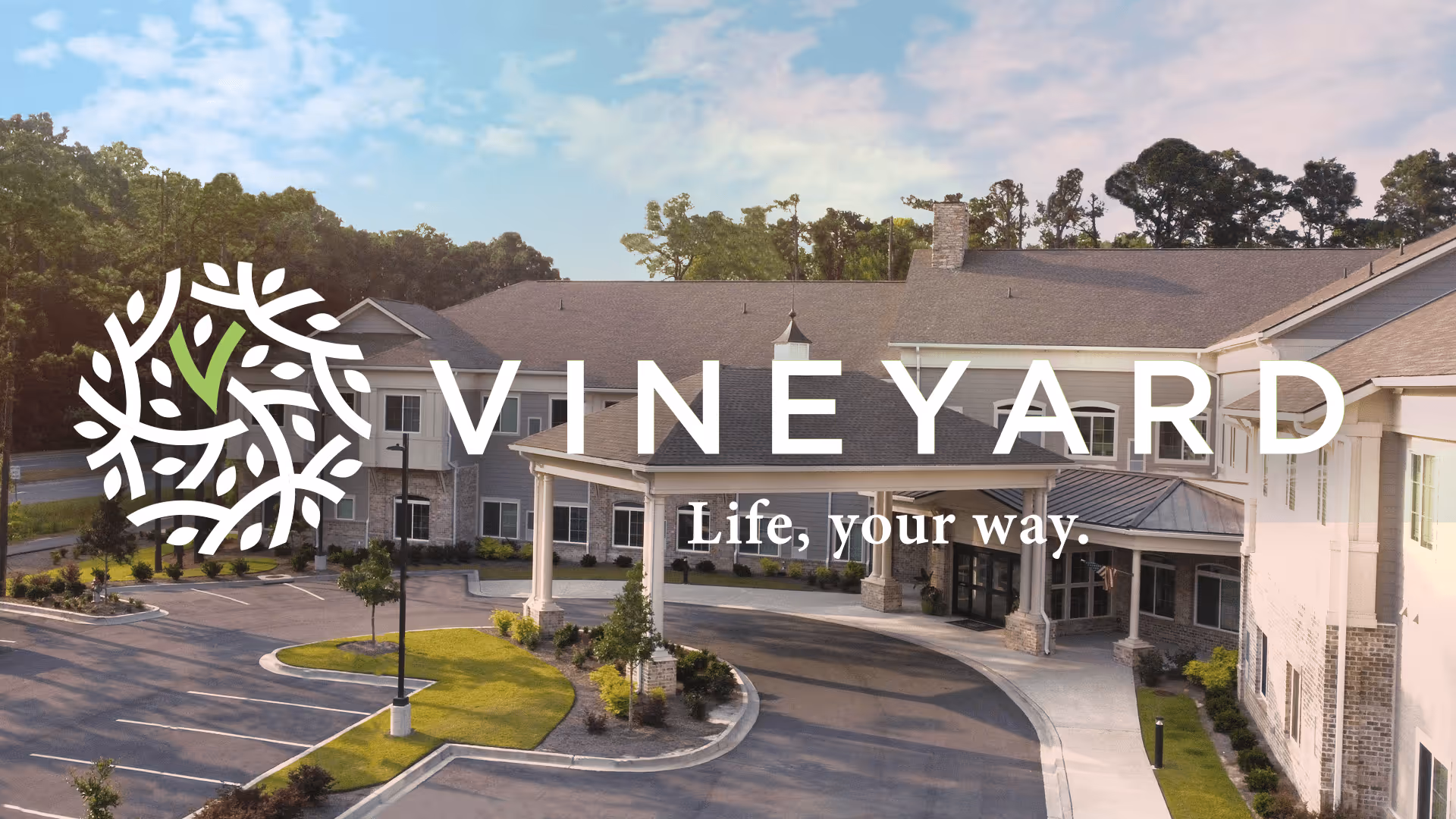 Exterior view of Vineyard Bluffton senior living facility showing a large building with a covered entrance, surrounded by landscaped greenery and a parking area under a partly cloudy sky.