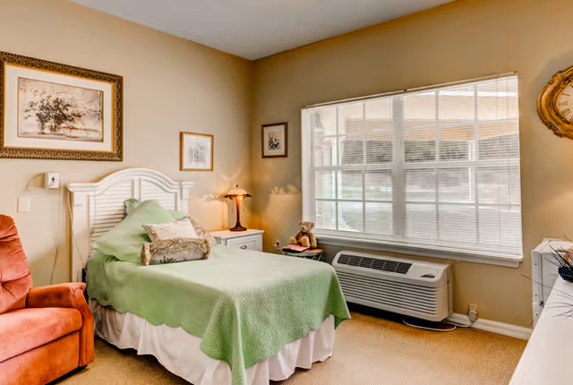 A cozy bedroom with a single bed draped in a green coverlet, bedside table and lamp, armchair, large window and wall art.