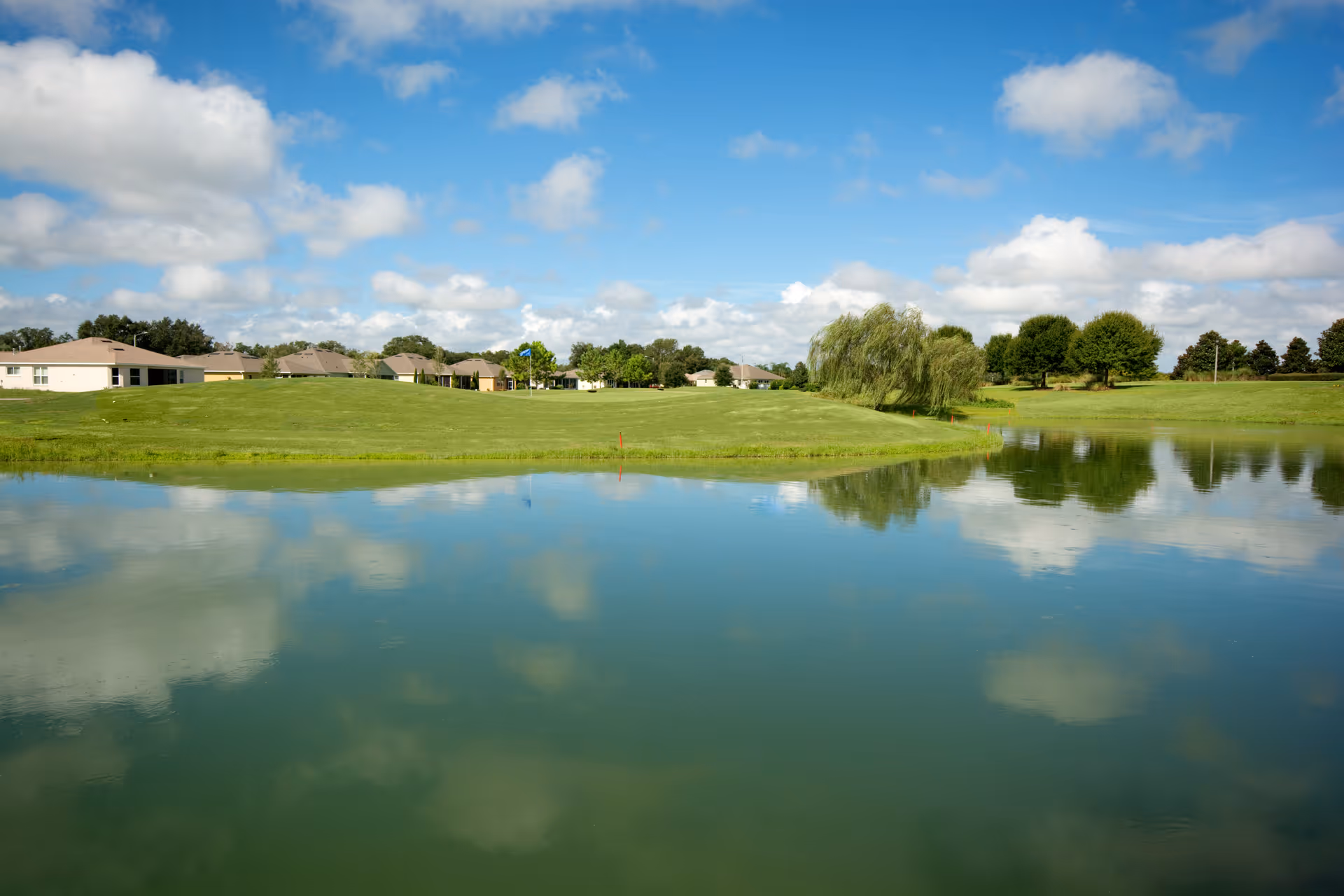 Calm pond reflecting clouds with a grassy landscape, trees, and single-story homes under a blue sky.