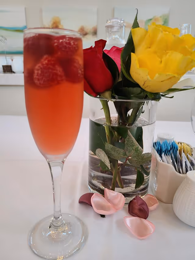 A close-up of a tall glass filled with a pinkish-red drink garnished with raspberries. Next to the glass is a clear vase with red and yellow roses and green leaves. Pink and burgundy rose petals are scattered on the white table surface. In the background, there is a container holding sugar and sweetener packets.