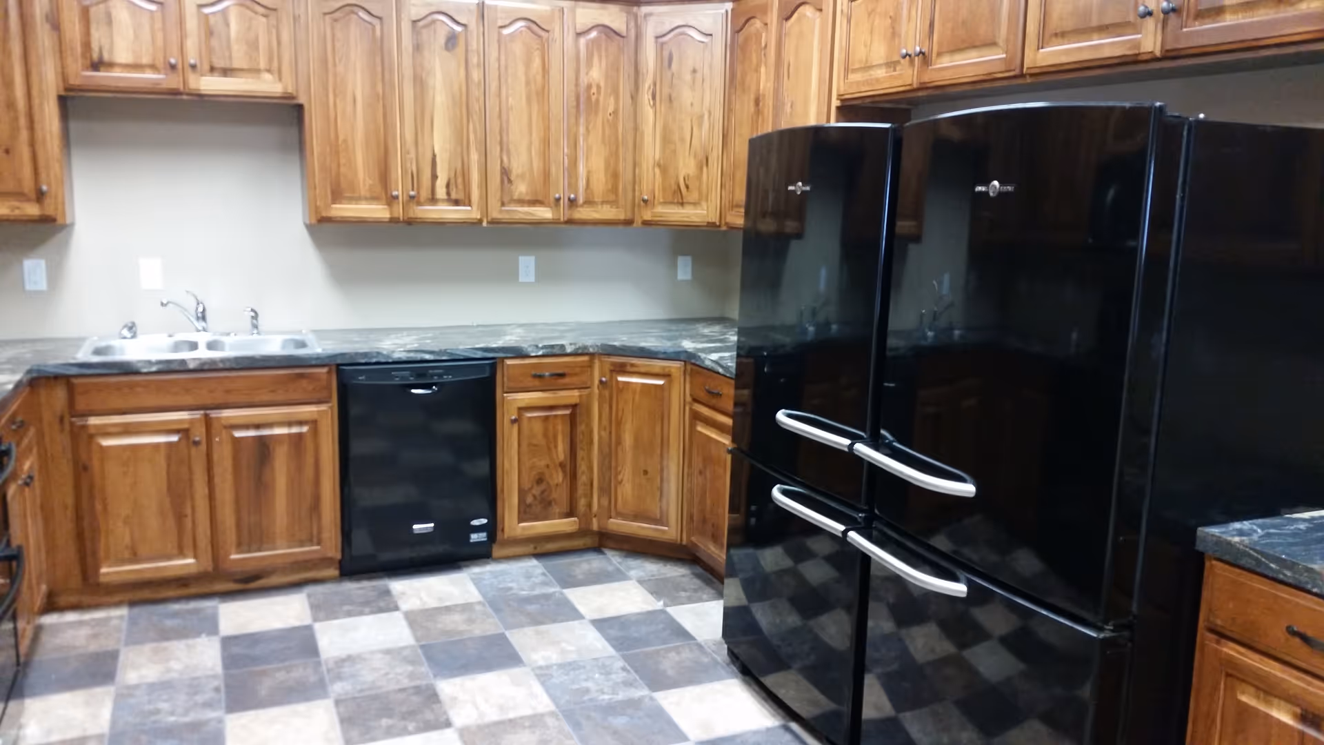 A kitchen with wooden cabinets, a double sink, a black dishwasher, and two black refrigerators. The floor has a checkered pattern with shades of brown and beige.