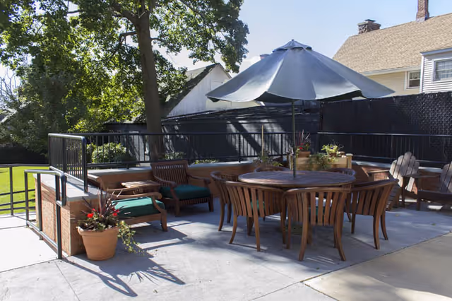 Outdoor patio area with a round wooden table surrounded by wooden chairs and a large umbrella providing shade. There are additional wooden chairs with green cushions, potted plants, and trees in the background. A building and a black fence are also visible.