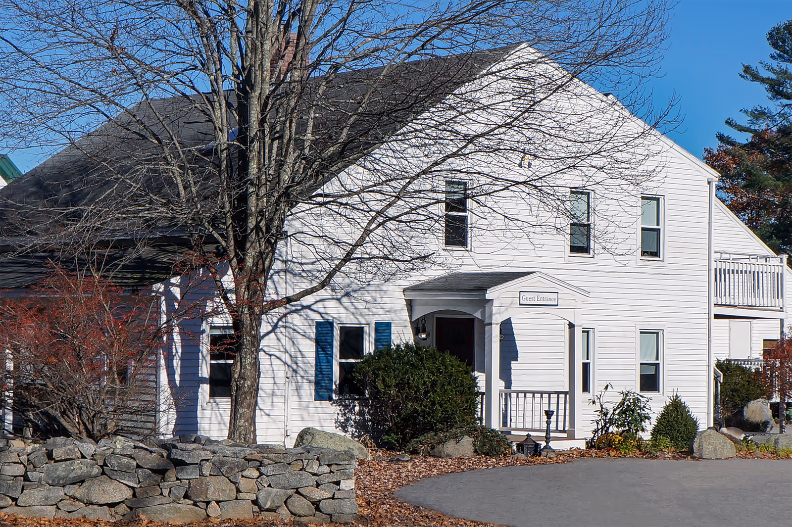 Exterior view of a white two-story building with a black roof, a stone wall in front, leafless trees, and a sign above the entrance that reads 'Guest Entrance'.