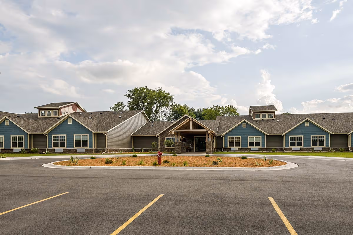 Front exterior view of Creamery Creek Senior Living Viroqua - Stoddard building with a circular driveway, landscaped center island, and a partly cloudy sky above.