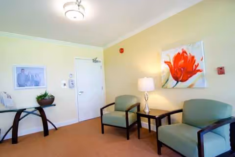 A small sitting area in a senior living facility with two green armchairs separated by a wooden side table holding a lamp. A large painting of a red flower hangs on the wall above the chairs. To the left, there is a glass-top console table with a plant and a framed photo on the wall above it. The walls are painted light yellow and the floor is carpeted in a light brown color.
