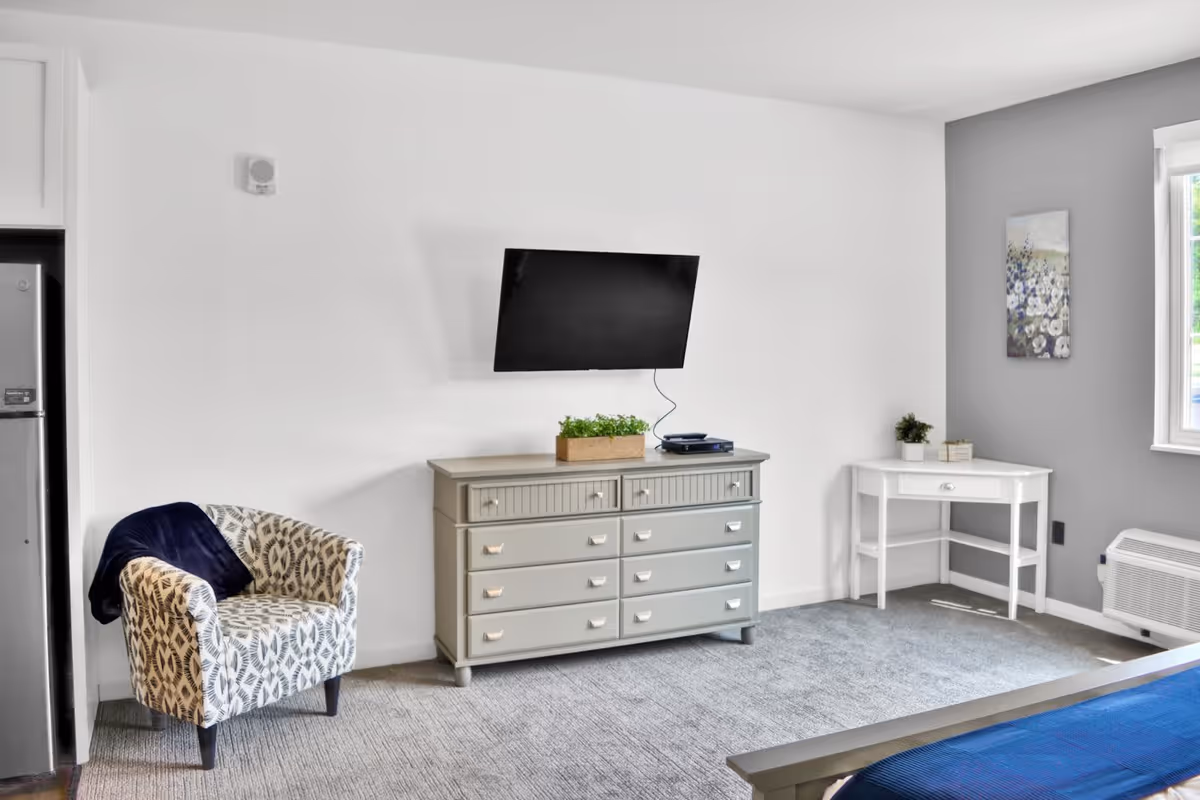 A bright and clean bedroom corner featuring a patterned armchair with a dark blue throw, a gray dresser with a small green plant and electronic device on top, a wall-mounted flat-screen TV, a white corner table with a small plant and books, a window with natural light, and part of a bed with a blue blanket.