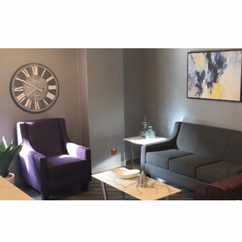 Cozy seating area with a purple armchair, gray sofa, coffee table, wall clock and artwork on the walls.