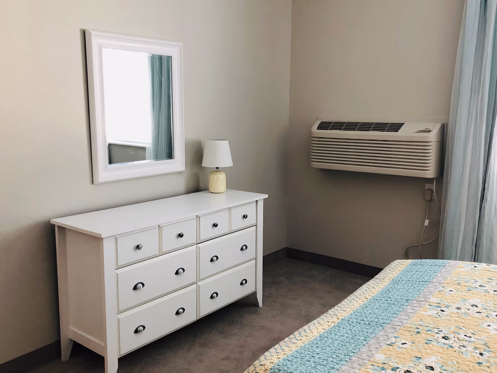 A bedroom corner featuring a white dresser with multiple drawers, a small lamp on top, and a white-framed mirror hanging above it. To the right, there is a wall-mounted air conditioning unit and a bed with a colorful quilted bedspread in shades of blue, yellow, and white. Light blue curtains hang by the window.