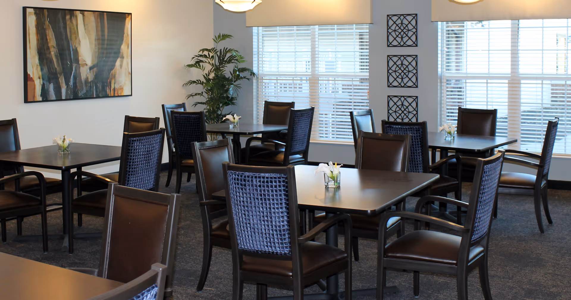A dining room with several dark wooden tables and chairs arranged neatly. Each table has a small flower vase with white flowers. The room has large windows with blinds, a potted plant in the corner, and abstract artwork on the wall.